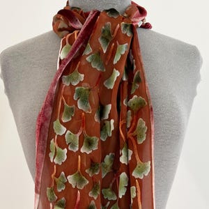 May include: A long, rectangular scarf featuring a sheer, rust-colored base fabric adorned with a repeating pattern of light green ginkgo leaves. The edges of the scarf are finished with a textured, velvet-like trim in a deep reddish-brown hue.