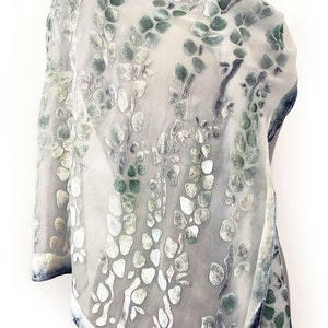 Puede incluir: Una bufanda blanca transparente con un estampado floral verde y blanco. La bufanda está hecha de una tela suave y ligera y tiene un borde con flecos.
