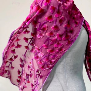 May include: A sheer, magenta scarf with a velvet floral pattern. The scarf is draped over a mannequin, showcasing its flowing design. The edges are trimmed with a darker magenta border, adding a touch of elegance to this accessory.