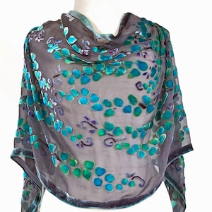 Puede incluir: Una bufanda gris transparente con un estampado floral en tonos turquesa, turquesa y morado. La bufanda tiene un ribete de terciopelo y está drapeada sobre un maniquí. El diseño floral está formado por pequeñas formas redondas.