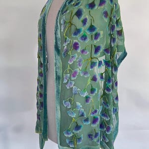 Op de afbeelding: Een doorschijnende, open groene sjaal met een fluwelen rand. De sjaal is versierd met een herhalend patroon van paarse en blauwe bloemmotieven. Het kledingstuk wordt op een mannequin getoond.