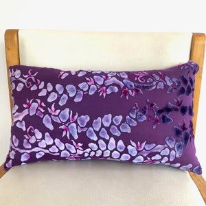 May include: Purple velvet pillow with a white floral pattern. The pillow is on a white chair with wooden arms.