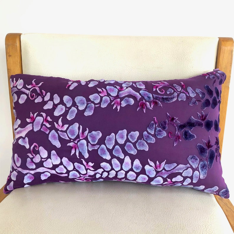 May include: Purple velvet pillow with a white floral pattern. The pillow is on a white chair with wooden arms.