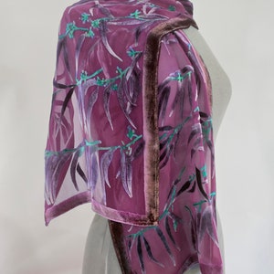 Puede incluir: Una bufanda de seda morada con un estampado floral y un ribete de terciopelo. La bufanda está drapeada sobre un maniquí.