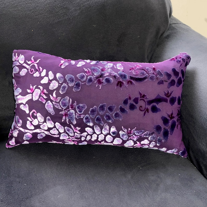 May include: Purple velvet pillow with a white floral pattern. The pillow is on a dark purple couch.