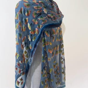May include: A blue semi-sheer scarf featuring a raised velvet ginkgo leaf pattern in shades of orange, yellow, and green. The scarf has a solid blue velvet border along the edges and is draped over a mannequin to display the texture and design.
