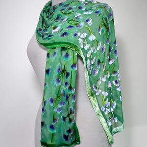 May include: A sheer green scarf with a floral pattern of blue, purple, and white flowers. The scarf has a velvet trim and is draped over a mannequin. The scarf is a light, airy accessory.