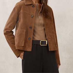 May include: A brown suede jacket with a button-up front and two front pockets. The jacket is paired with a brown henley shirt, black pants, and a black belt with a gold buckle. The jacket is a classic style.