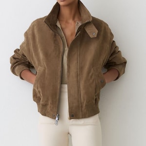 May include: A brown suede bomber jacket with a high collar and a zipper closure. The jacket has ribbed cuffs and a ribbed hem. The model is wearing a beige top and cream-colored pants. The jacket has two front pockets.
