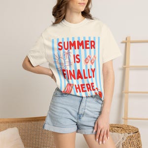 Puede incluir: Camiseta color crema con el texto "SUMMER IS FINALLY HERE" en rojo y azul. El diseño incluye un sol, palmeras y chanclas. La camiseta se combina con pantalones cortos vaqueros.