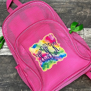 May include: A bright pink mesh backpack featuring a central patch with a colorful tie-dye pattern and dark purple cursive monogram lettering. The backpack includes a top handle, a front zippered pocket, and small green ribbon accents on the zippers.