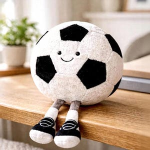 Könnte beinhalten: Ein Stofffußball mit einem lächelnden Gesicht, schwarz-weißen Paneelen und angebrachten Beinen mit schwarz-weißen Turnschuhen. Das Plüschtier ist für Kinder konzipiert und sitzt auf einer Holzoberfläche.