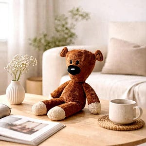 May include: A brown teddy bear plush toy with a black nose and cream-colored paws and feet. The bear is sitting on a wooden table next to a white mug and a woven coaster. A vase of white flowers and an open book are also on the table.