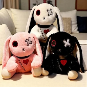 May include: Three plush bunny toys in white, pink, and black. Each bunny has long ears, a stitched cross on one eye, and a red heart on the chest. The bunnies have a gothic aesthetic.
