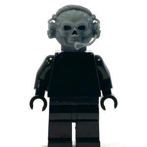 Könnte beinhalten: Eine schwarze Lego-Figur mit grauem Totenkopf, die ein Headset mit Mikrofon trägt. Die Figur hat schwarze Arme und Beine und steht vor einem weißen Hintergrund.