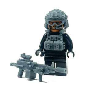 Op de afbeelding: Een grijze Lego-figuur met militair thema, met helm, tactisch vest en machinegeweer. De figuur is zwart en grijs, met een gedetailleerd ontwerp. Het machinegeweer heeft een vizier en andere opzetstukken. De figuur staat op een witte achtergrond.