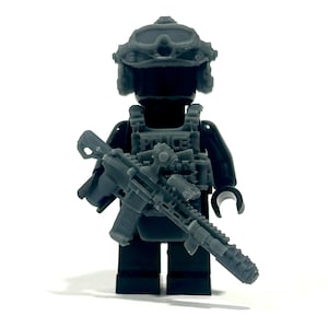 Op de afbeelding: Een grijze Lego-figuur van een politieagent in tactische uitrusting, inclusief helm, vest met het woord "POLICE" en een geweer. De figuur is zwart met grijze accessoires.