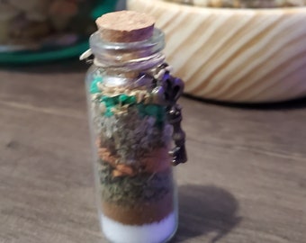 May include: A small glass bottle with a cork stopper filled with layers of dried herbs and spices. The bottle has a metal charm attached to it.