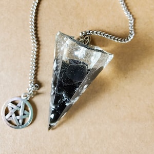 May include: A clear, triangular pendulum with black and white inclusions, suspended by a silver chain. A silver pentagram charm hangs from the chain. The pendulum is likely used for divination or spiritual practices.