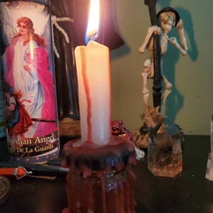 May include: A white candle with red wax dripping down the sides is burning in a glass jar with a black lid. The candle is surrounded by other objects, including a crystal, a statue of a skeleton, and a religious candle.