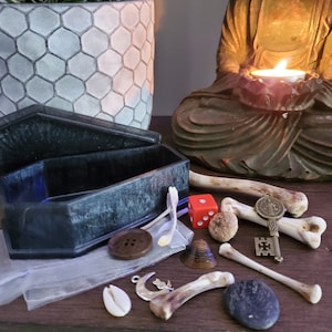 May include: A black coffin-shaped box with a blue interior, a red dice, a small wooden button, a seashell, a key with a charm, a bone, a small stone, and a wooden object that looks like a small cone.