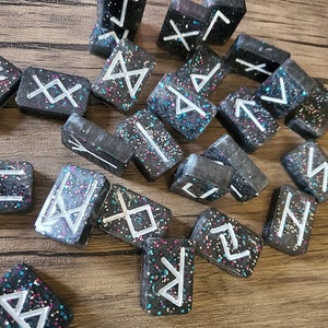 May include: A set of black and white rune stones with silver glitter. The runes are painted in white on the black stones.