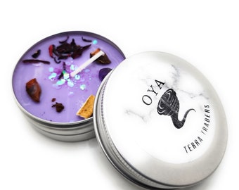 May include: A purple candle with glitter and dried flowers in a silver tin. The lid of the tin has a black and white marble design with the text "OYA TERRA TRADERS" and a black swirl design.