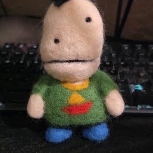 May include: A needle-felted doll with a cream-colored head, a small black tuft of hair, and two black dot eyes. The doll wears a green shirt featuring a yellow triangle and a red crescent shape, paired with bright blue pants.
