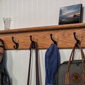 May include: A horizontal wooden wall-mounted coat rack featuring four black metal hooks. The rack has a flat top shelf surface. Items hanging from the hooks include a plaid cap, a dark strap, a blue fabric item, and a gray backpack with brown handles.
