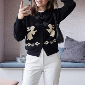 Peut inclure: Cardigan noir avec fermetures à bascule et motif de lapin et floral crème. Le pull est porté avec un pantalon blanc. Un sac à main noir est visible.