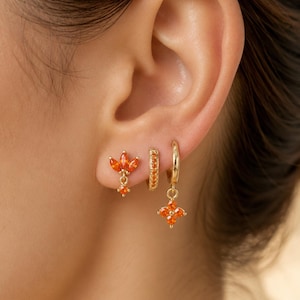 May include: A set of three gold-toned earrings featuring orange gemstones. The first is a stud with three marquise-cut stones and a dangling round stone. The second is a small hoop with channel-set stones. The third is a hoop with a flower charm.