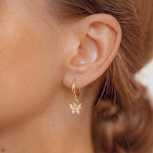 Puede incluir: Un pendiente de aro dorado con un pequeño dije de mariposa. El dije de mariposa está incrustado con pequeños cristales transparentes. El pendiente se muestra en la oreja de una persona, centrándose en la joya.