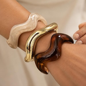 Peut inclure: Un ensemble de trois bracelets rigides ondulés portés au poignet. Le premier bracelet est en résine blanc cassé, le deuxième est en métal doré poli et le troisième est en résine brun foncé translucide. Chaque bracelet présente une forme ondulée.
