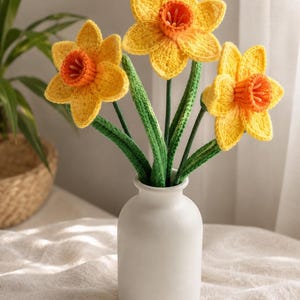 May include: Three crocheted daffodil flowers with bright yellow petals and orange centers, attached to long green stems with leaves, arranged in a simple white ceramic vase with a narrow neck and a rounded base.