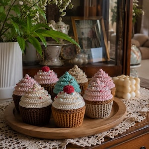 Può includere: Una collezione di cupcake all'uncinetto fatti a mano in vari colori, tra cui rosa, bianco, marrone e verde acqua, ognuno sormontato da una ciliegia all'uncinetto. I cupcake sono disposti su un vassoio di legno, mostrando dettagli e trame.