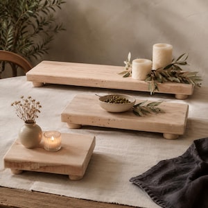 May include: Three rectangular light-toned wooden risers with short, rounded feet. The largest riser holds two pillar candles and olive branches. The medium riser holds a small bowl of green olives. The smallest riser holds a vase and candle.