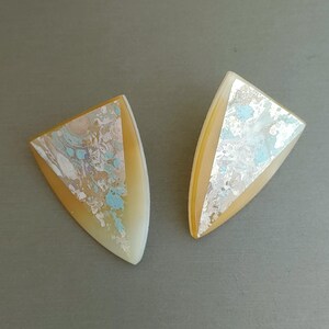May include: Two abstract earrings with a shield shape. The earrings have a light yellow border and a center with a mix of colors, including white, blue, and silver. The earrings are set against a gray background.