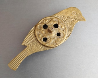 May include: A brass bird-shaped object with a circular, patterned centerpiece. The bird's body features etched details, and the circular element has four holes. The object has a vintage aesthetic.