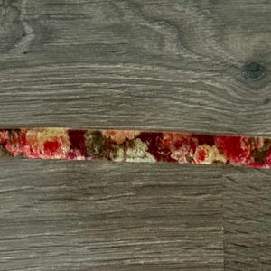 May include: A narrow strip of velvet ribbon featuring a repeating floral pattern in shades of deep red, muted gold, and dark brown. The fabric has a soft, textured pile and is displayed horizontally across the frame in a single continuous line.