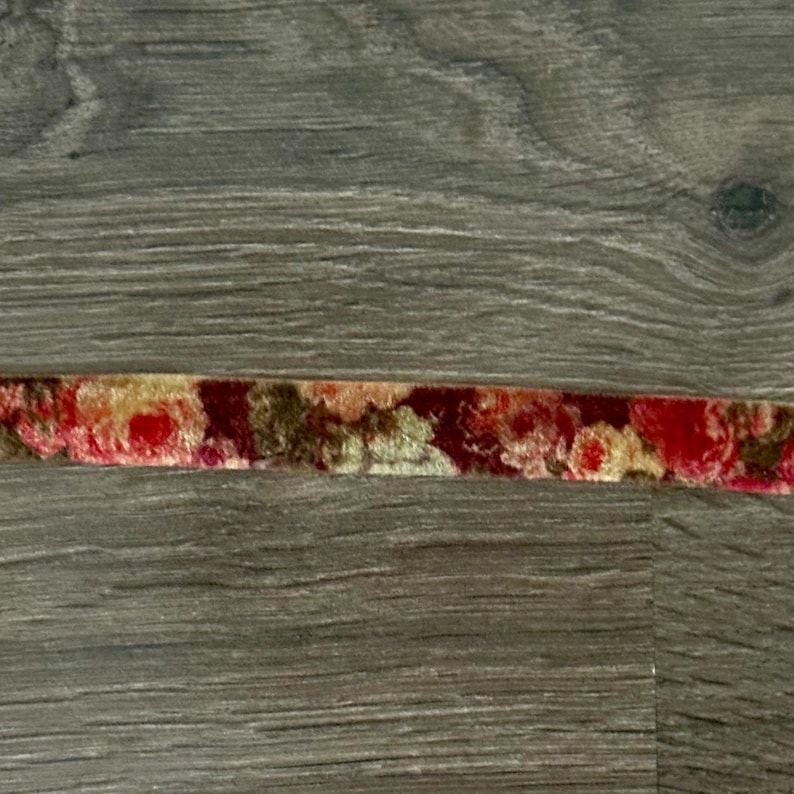 May include: A narrow strip of velvet ribbon featuring a repeating floral pattern in shades of deep red, muted gold, and dark brown. The fabric has a soft, textured pile and is displayed horizontally across the frame in a single continuous line.