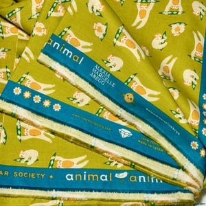 May include: A folded piece of olive green cotton fabric featuring a repeating pattern of white sloths hanging from green patterned branches. The selvage edge is teal with white text reading animal, Alexia Marcelle Abegg, and Ruby Star Society.