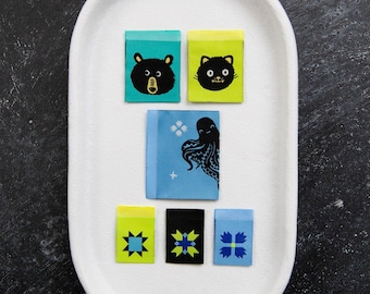 May include: Six rectangular fabric labels featuring various designs: a black bear face on teal, a black cat face on lime green, an octopus on light blue, and three smaller labels with geometric star patterns in yellow, black, and light blue.