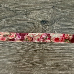 May include: A strip of velvet ribbon featuring a repeating floral pattern in shades of deep red, bright pink, and muted green. The fabric has a soft, textured pile and displays a dense arrangement of roses and leaves across its entire length.