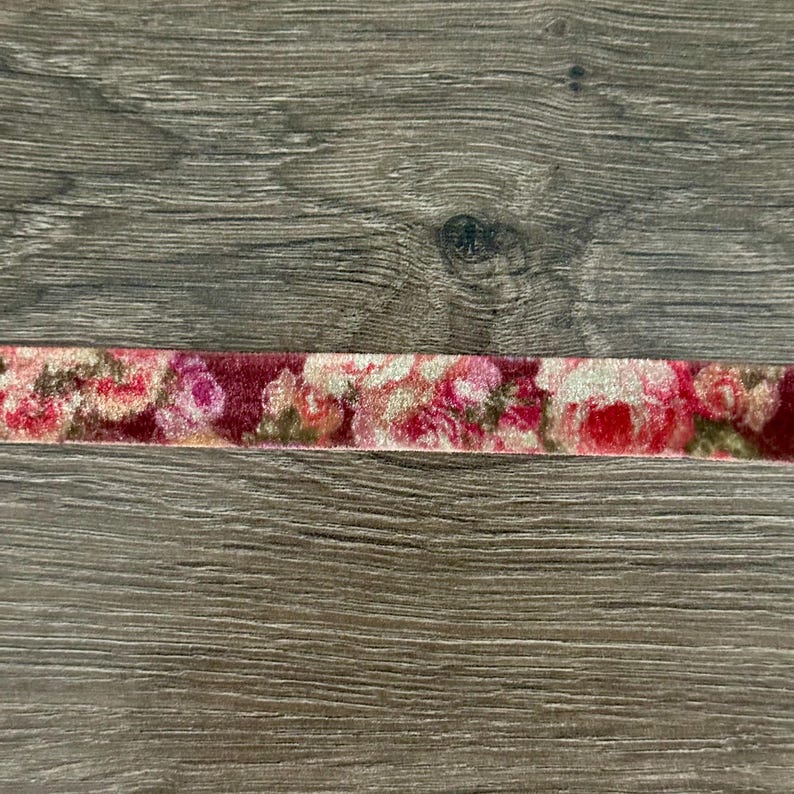 May include: A strip of velvet ribbon featuring a repeating floral pattern in shades of deep red, bright pink, and muted green. The fabric has a soft, textured pile and displays a dense arrangement of roses and leaves across its entire length.