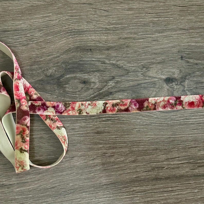 May include: A length of velvet ribbon featuring a floral pattern in shades of pink, red, and cream. The ribbon has a soft, textured surface with a repeating rose print and a plain, light-colored backing visible on the underside of the loops.