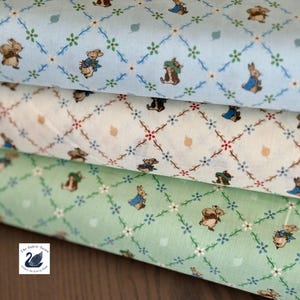May include: Three stacked rolls of fabric featuring a repeating pattern of small brown rabbits and floral diamond lattice designs. The top roll is light blue, the middle is off-white, and the bottom is light green. A small logo with a swan is visible.