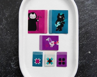 May include: A collection of six rectangular fabric labels featuring graphic designs including a cat in a black robe with a scythe, a bear playing a guitar, a dachshund, and three labels with geometric star patterns in pink, blue, and purple.