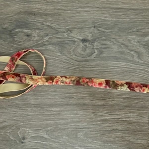 May include: A length of velvet ribbon featuring a floral pattern in shades of red, pink, and cream on the front side, with a solid beige backing. The ribbon is coiled at one end and extends horizontally across the frame in a straight line.