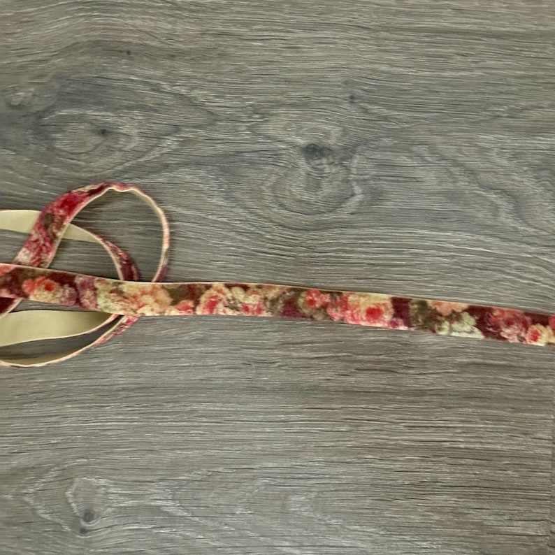 May include: A length of velvet ribbon featuring a floral pattern in shades of red, pink, and cream on the front side, with a solid beige backing. The ribbon is coiled at one end and extends horizontally across the frame in a straight line.