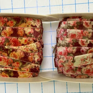 May include: Two spools of velvet ribbon featuring a floral pattern with shades of deep red, burgundy, and muted yellow. The ribbon is wound tightly around circular plastic spools, displaying a repeating rose print across the soft fabric surface.